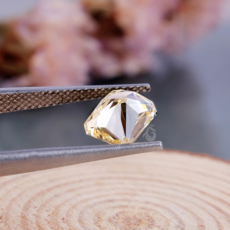 May include: A faceted, pale yellow gemstone held by silver-toned metal tweezers. The stone features a geometric cut with multiple reflective surfaces that catch the light, displaying a clear, translucent quality and a distinct, sharp-edged shape.
