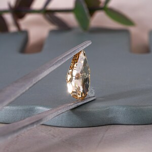 May include: A pear-shaped, champagne-colored gemstone held by metal tweezers. The stone features multiple facets that reflect light, creating a brilliant, sparkling appearance. The gem is transparent with a warm, brownish-gold hue and a pointed top.