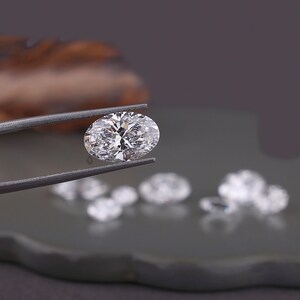 May include: A clear, oval-cut gemstone with multiple facets held by dark metal tweezers. The stone displays high brilliance and light reflection. Several other smaller, out-of-focus, clear, faceted gemstones are positioned in the background.