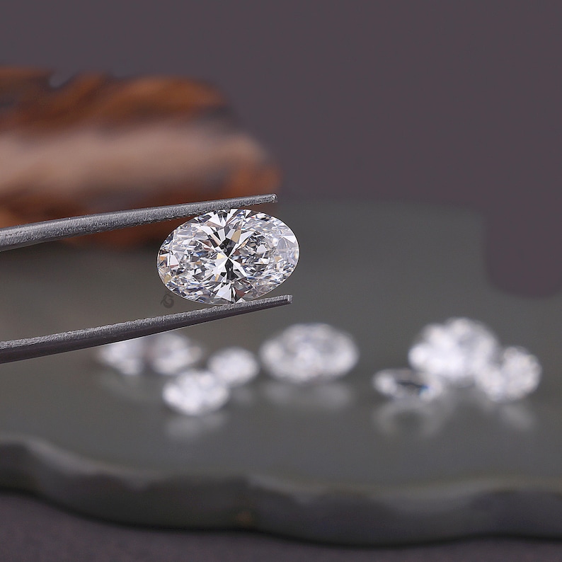 May include: A clear, oval-cut gemstone with multiple facets held by dark metal tweezers. The stone displays high brilliance and light reflection. Several other smaller, out-of-focus, clear, faceted gemstones are positioned in the background.
