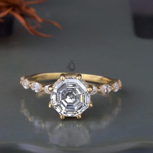 May include: A gold ring featuring a large, octagonal-cut diamond as the centerpiece. The band is adorned with smaller, marquise-cut diamonds. The ring is set against a neutral background, highlighting its sparkle and intricate design.