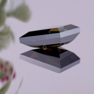 May include: A dark, faceted gemstone with a rectangular, emerald-cut shape. The stone displays deep charcoal and black tones with sharp, geometric edges and a polished, reflective surface. A small, gold-coloured crown logo is visible on the stone.