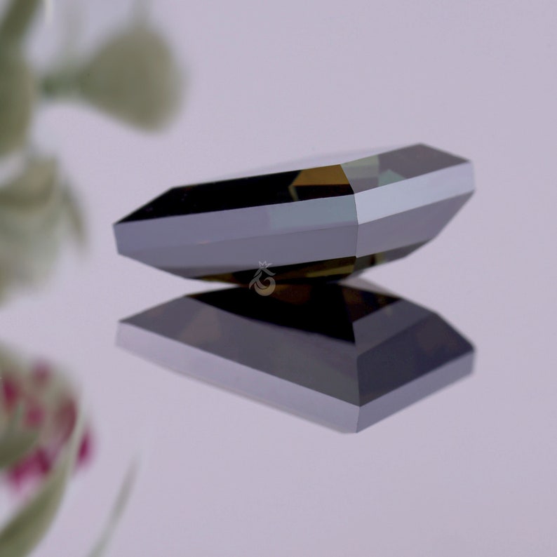 May include: A dark, faceted gemstone with a rectangular, emerald-cut shape. The stone displays deep charcoal and black tones with sharp, geometric edges and a polished, reflective surface. A small, gold-coloured crown logo is visible on the stone.