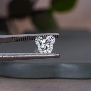 May include: A clear, heart-shaped diamond with a unique, rounded, Mickey Mouse-like design. The diamond is held in tweezers.