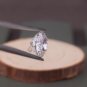 May include: A marquise-cut, colorless, faceted gemstone held by dark metal tweezers. The stone features a brilliant, multi-faceted surface that reflects light, creating a clear, sparkling appearance with sharp, pointed ends and a curved center.