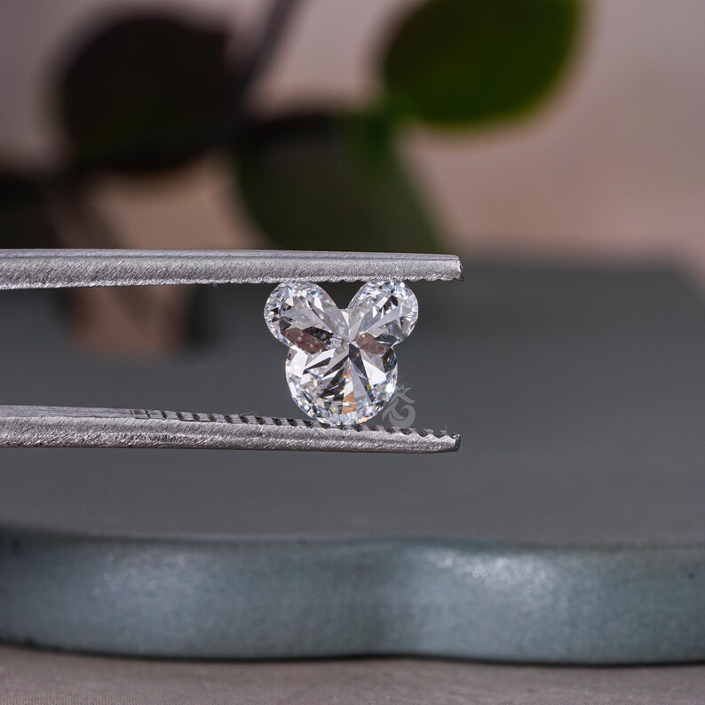 May include: A clear, faceted, heart-shaped diamond with a Mickey Mouse design. The diamond is held in tweezers.