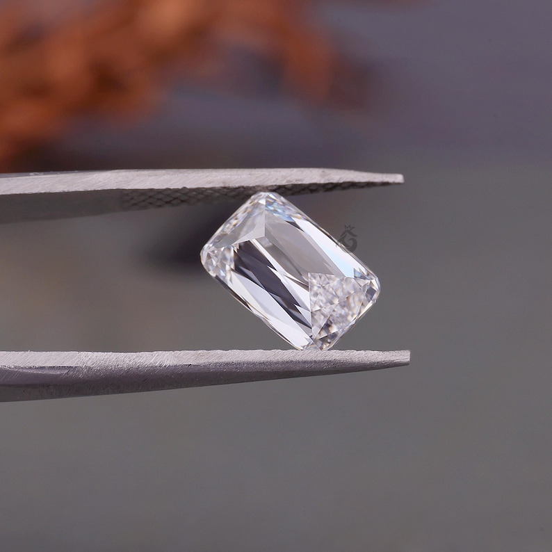 May include: A clear, rectangular, emerald-cut gemstone held by metal tweezers. The stone features multiple facets that reflect light, creating bright white highlights and dark shadows within its transparent, crystalline structure.