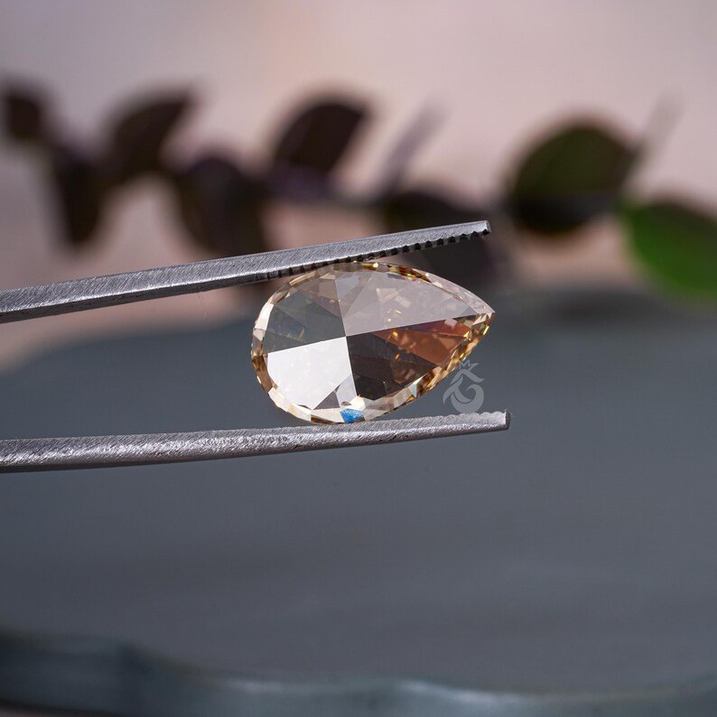 May include: A pear-shaped, champagne-colored gemstone held by a pair of metal tweezers. The stone features a faceted cut that reflects light, creating distinct geometric patterns across its surface. A small logo is visible on the stone's face.