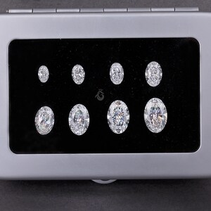 May include: A rectangular silver-toned storage case with a black velvet interior containing eight clear, oval-cut faceted gemstones arranged in two rows of four, with a small crown-shaped logo printed in the center of the black display surface.