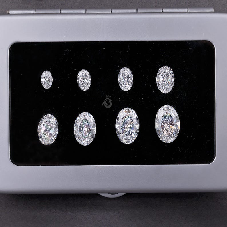 May include: A rectangular silver-toned storage case with a black velvet interior containing eight clear, oval-cut faceted gemstones arranged in two rows of four, with a small crown-shaped logo printed in the center of the black display surface.