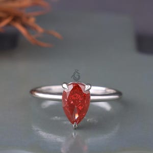 May include: A silver ring featuring a pear-shaped, vibrant red gemstone. The gemstone is the focal point, set with four prongs on a thin, polished band. The ring is displayed on a neutral gray surface.