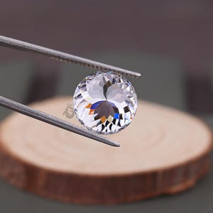 May include: A round, clear, faceted gemstone held by metal tweezers. The stone features a brilliant cut with numerous triangular facets that reflect light, creating a sparkling appearance. A small logo is visible on the surface of the gemstone.