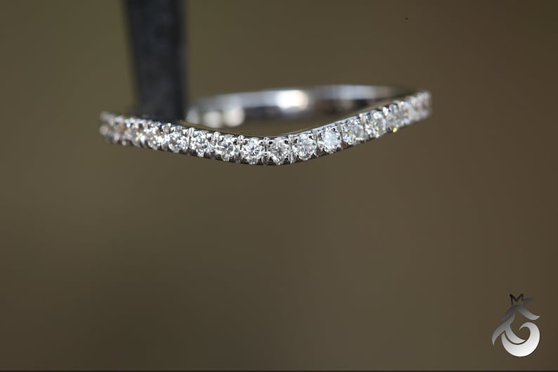 May include: A silver-toned curved band ring featuring a row of small, round, clear faceted stones set along the top edge. The metal has a polished, reflective finish, and the design follows a gentle V-shape contour, showcasing multiple stones.