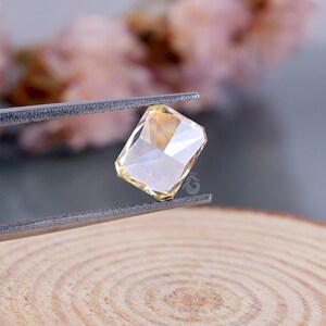 May include: A rectangular, faceted, pale yellow gemstone held by silver-toned metal tweezers. The stone features a clear, brilliant cut with multiple reflective surfaces that catch the light, displaying a soft, warm, translucent golden hue.