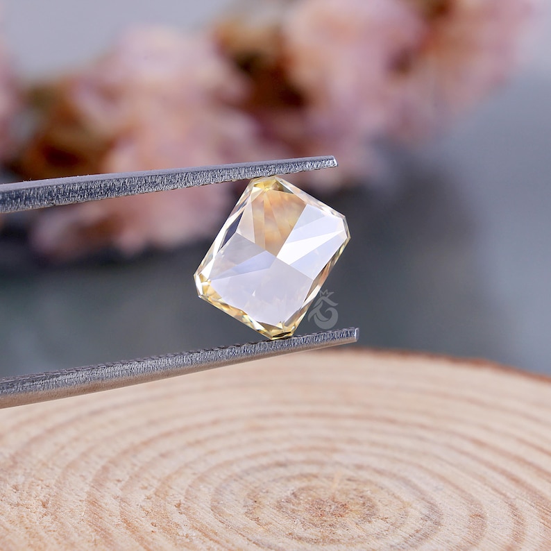 May include: A rectangular, faceted, pale yellow gemstone held by silver-toned metal tweezers. The stone features a clear, brilliant cut with multiple reflective surfaces that catch the light, displaying a soft, warm, translucent golden hue.