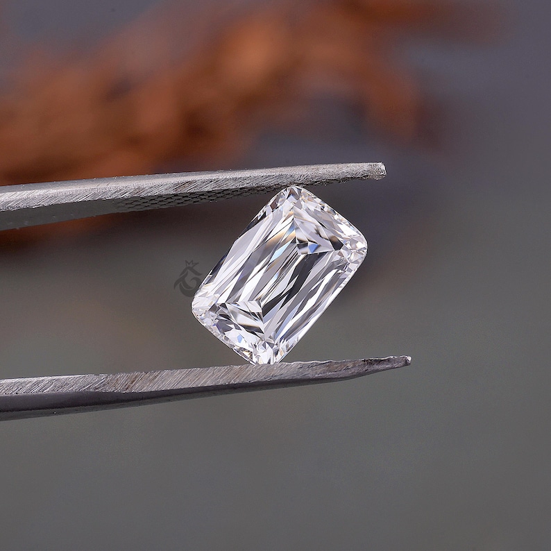 May include: A clear, colourless, rectangular emerald-cut gemstone held by metal tweezers. The stone features sharp, geometric facets that reflect light, creating a brilliant, multi-faceted appearance with a small logo mark visible on the surface.
