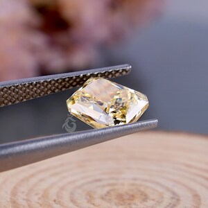May include: A radiant-cut yellow gemstone held by metal tweezers. The stone features a faceted surface that reflects light, creating a bright, translucent appearance with a pale golden hue. A small logo is visible on the side of the gemstone.