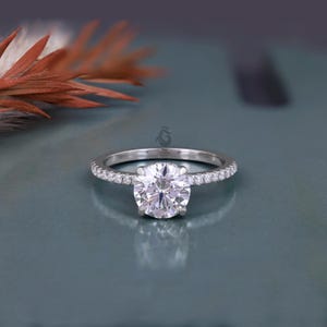 Puede incluir: Un anillo de tono plateado con una gran piedra central redonda de talla brillante sujeta por cuatro garras. La banda es delgada y está adornada con una hilera de pequeñas piedras redondas engastadas en pavé a lo largo de la mitad superior.