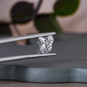 May include: A clear heart-shaped diamond gemstone held in a pair of silver tweezers. The diamond is set against a gray background.