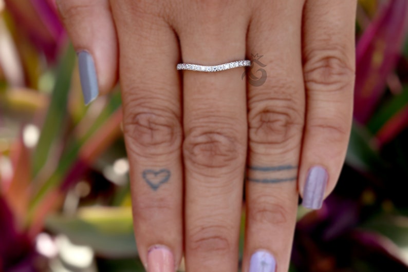 May include: A curved silver-toned band ring featuring a row of small, clear, sparkling stones set along the top surface. The ring has a distinct V-shaped or chevron contour, designed to sit flush against another ring on the finger.
