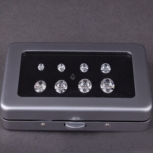 May include: A rectangular gray metal storage case with a clear window lid containing eight round, faceted, clear gemstones arranged in two rows of four on a black velvet insert. A small logo is printed between the two rows of stones.