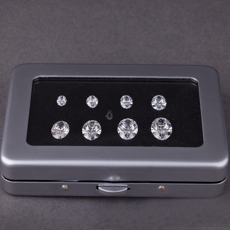 May include: A rectangular gray metal storage case with a clear window lid containing eight round, faceted, clear gemstones arranged in two rows of four on a black velvet insert. A small logo is printed between the two rows of stones.