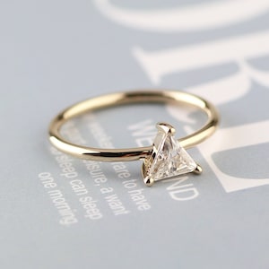 May include: A gold ring featuring a single, clear, triangular-cut gemstone set in a three-prong setting. The band is thin, smooth, and polished, showcasing a minimalist design that highlights the geometric shape of the central stone.