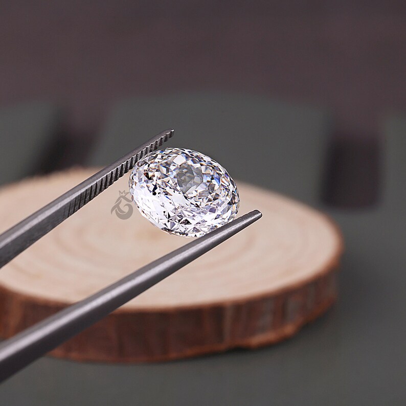 May include: A clear, faceted oval gemstone held by metal tweezers. The stone features a brilliant cut with numerous reflective surfaces that catch the light, creating a sparkling appearance. A small logo is visible on the side of the gemstone.