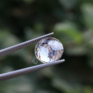 May include: A round, faceted, clear gemstone held by metal tweezers. The stone features a brilliant cut with numerous triangular facets that reflect light, creating a sparkling appearance. A small logo is visible on the surface of the stone.