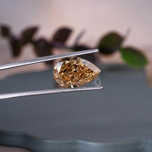 May include: A pear-shaped, faceted gemstone with a warm, golden-brown hue held by a pair of silver-toned metal tweezers. The stone exhibits multiple reflective surfaces and internal light refraction, showcasing a clear, brilliant, and polished finish.