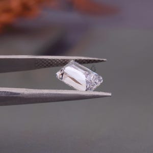 May include: A clear, rectangular-cut gemstone held by metal tweezers. The stone features multiple facets that reflect light, creating bright white highlights and internal shadows. A small, faint logo is visible on the surface of the gemstone.
