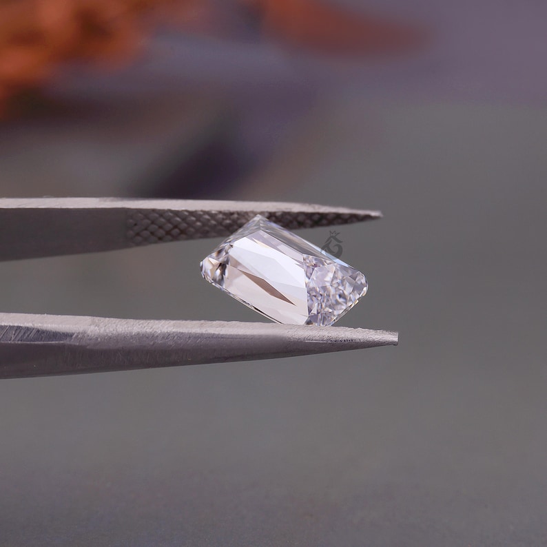 May include: A clear, rectangular-cut gemstone held by metal tweezers. The stone features multiple facets that reflect light, creating bright white highlights and internal shadows. A small, faint logo is visible on the surface of the gemstone.