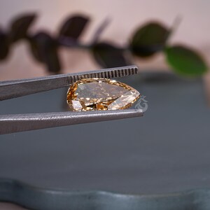 May include: A pear-shaped, cognac-colored gemstone held by a pair of metal tweezers. The faceted stone exhibits a warm, golden-brown hue with internal reflections and a clear, polished surface. A small, faint logo is visible on the stone's surface.