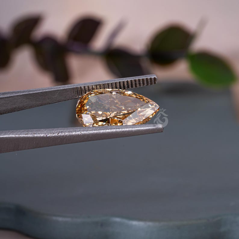 May include: A pear-shaped, cognac-colored gemstone held by a pair of metal tweezers. The faceted stone exhibits a warm, golden-brown hue with internal reflections and a clear, polished surface. A small, faint logo is visible on the stone's surface.