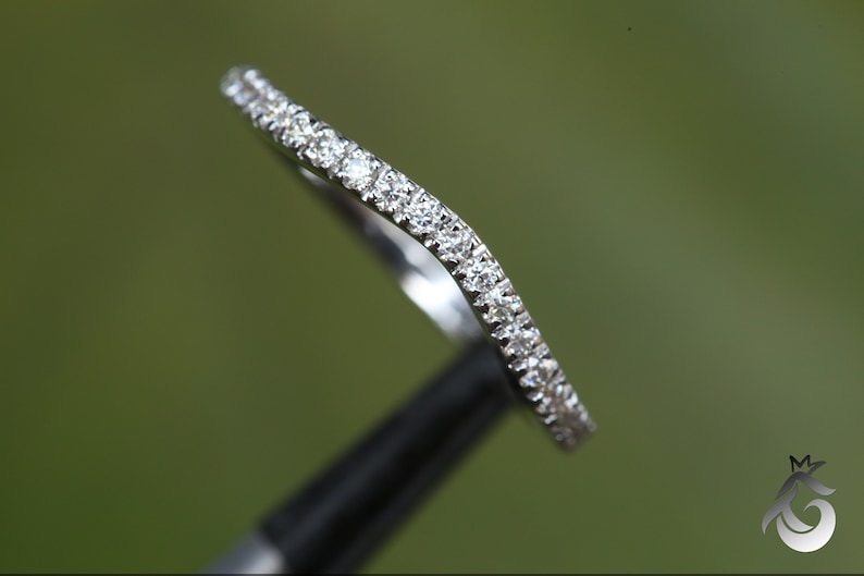 May include: A curved silver-toned ring band featuring a row of small, clear, round-cut stones set along the top surface. The metal has a polished, reflective finish, and the band follows a gentle wave-like contour throughout its entire length.