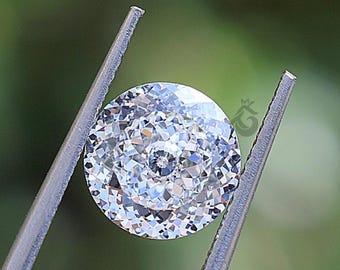 May include: A round, brilliant-cut, colourless gemstone with numerous triangular and kite-shaped facets that create a high level of light reflection and sparkle. The stone is held by metal tweezers and features a small, crown-shaped logo mark.