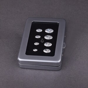 May include: A rectangular gray metal display case with a clear window lid, containing eight oval-cut clear gemstones arranged in two vertical columns on a black velvet insert. The stones vary in size, with the largest positioned at the top right.