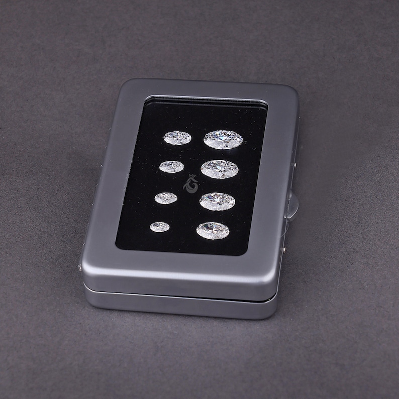 May include: A rectangular gray metal display case with a clear window lid, containing eight oval-cut clear gemstones arranged in two vertical columns on a black velvet insert. The stones vary in size, with the largest positioned at the top right.