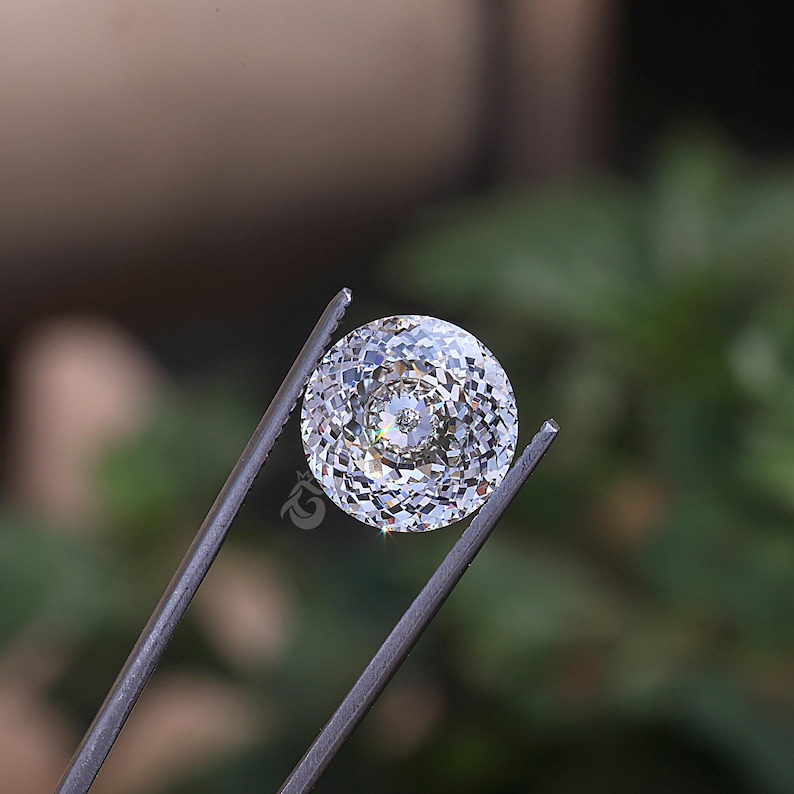 May include: A round, colorless, faceted gemstone with a brilliant cut, displaying multiple triangular and kite-shaped facets that reflect light. The stone is held by metal tweezers and features a small logo mark on its lower left side.