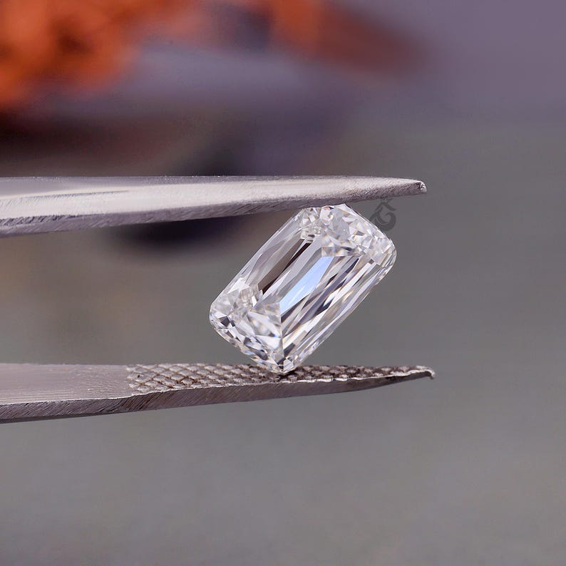 May include: A clear, rectangular emerald-cut gemstone held by metal tweezers. The stone features multiple facets that reflect light, creating a brilliant, colourless appearance with sharp, clean edges and a high degree of transparency and clarity.