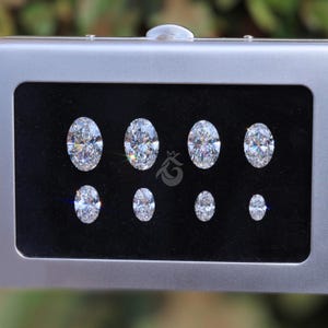 May include: A silver-toned rectangular display case containing eight oval-cut clear gemstones arranged in two rows on a black velvet insert. The stones vary in size, and a small logo featuring a stylized letter G with a crown is visible in the center.