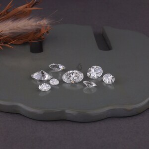 May include: A collection of eight loose, clear, faceted gemstones of varying sizes and shapes, including oval and round cuts, arranged on a smooth, dark gray surface. The stones exhibit high clarity and brilliance, reflecting light from multiple angles.