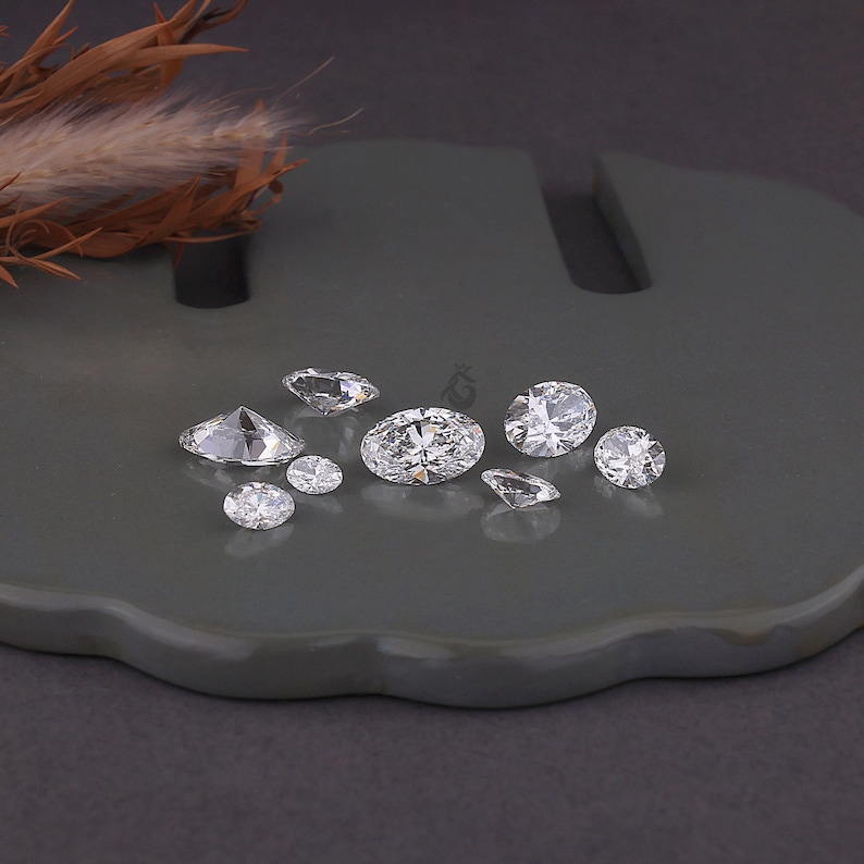 May include: A collection of eight loose, clear, faceted gemstones of varying sizes and shapes, including oval and round cuts, arranged on a smooth, dark gray surface. The stones exhibit high clarity and brilliance, reflecting light from multiple angles.
