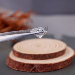 May include: A clear, faceted, heart-shaped gemstone held by silver-toned metal tweezers. The stone features multiple triangular and kite-shaped facets that reflect light, creating a brilliant, transparent appearance with sharp, defined edges.