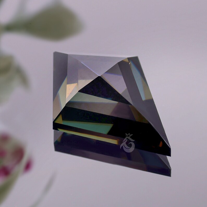 May include: A kite-shaped gemstone with a faceted surface displaying dark blue, green, and purple iridescent tones. The stone features a small, etched crown-shaped logo on its lower right facet, reflecting light with sharp geometric edges.