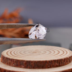 May include: A clear, faceted, cushion-cut gemstone held by metal tweezers. The stone features multiple triangular and trapezoidal facets that reflect light, creating a brilliant, transparent appearance. A small logo is visible on the stone surface.