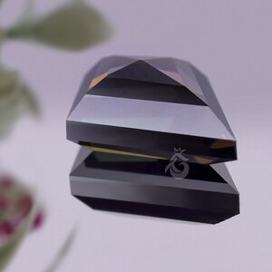 May include: A faceted, dark grey gemstone with a rectangular, step-cut design. The stone features a sharp, pyramid-like top and a flat base, reflecting light with a metallic sheen. A small, white crown-shaped logo is visible on the side.