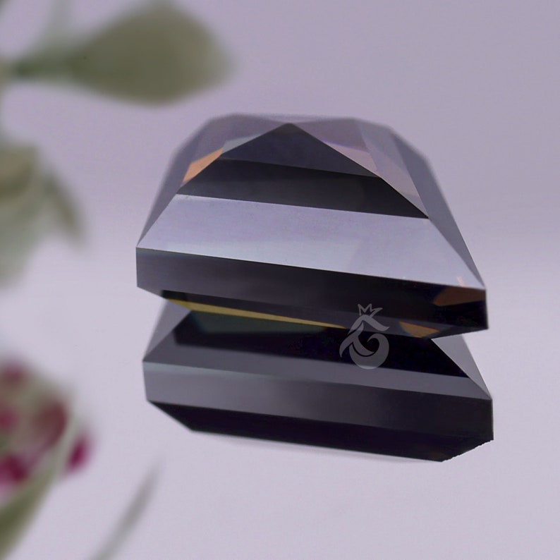 May include: A faceted, dark grey gemstone with a rectangular, step-cut design. The stone features a sharp, pyramid-like top and a flat base, reflecting light with a metallic sheen. A small, white crown-shaped logo is visible on the side.
