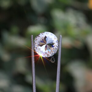 May include: A round, clear, faceted gemstone held by metal tweezers. The stone features a complex, star-like internal pattern with sharp, geometric cuts that reflect light, creating a bright, multi-colored flare at the lower left edge.