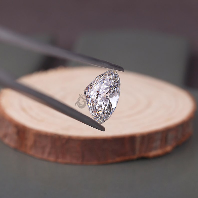 May include: A clear, pear-shaped faceted gemstone held by dark metal tweezers. The stone features numerous triangular and kite-shaped facets that reflect light, creating a brilliant, sparkling appearance with a distinct pointed end and a rounded top.